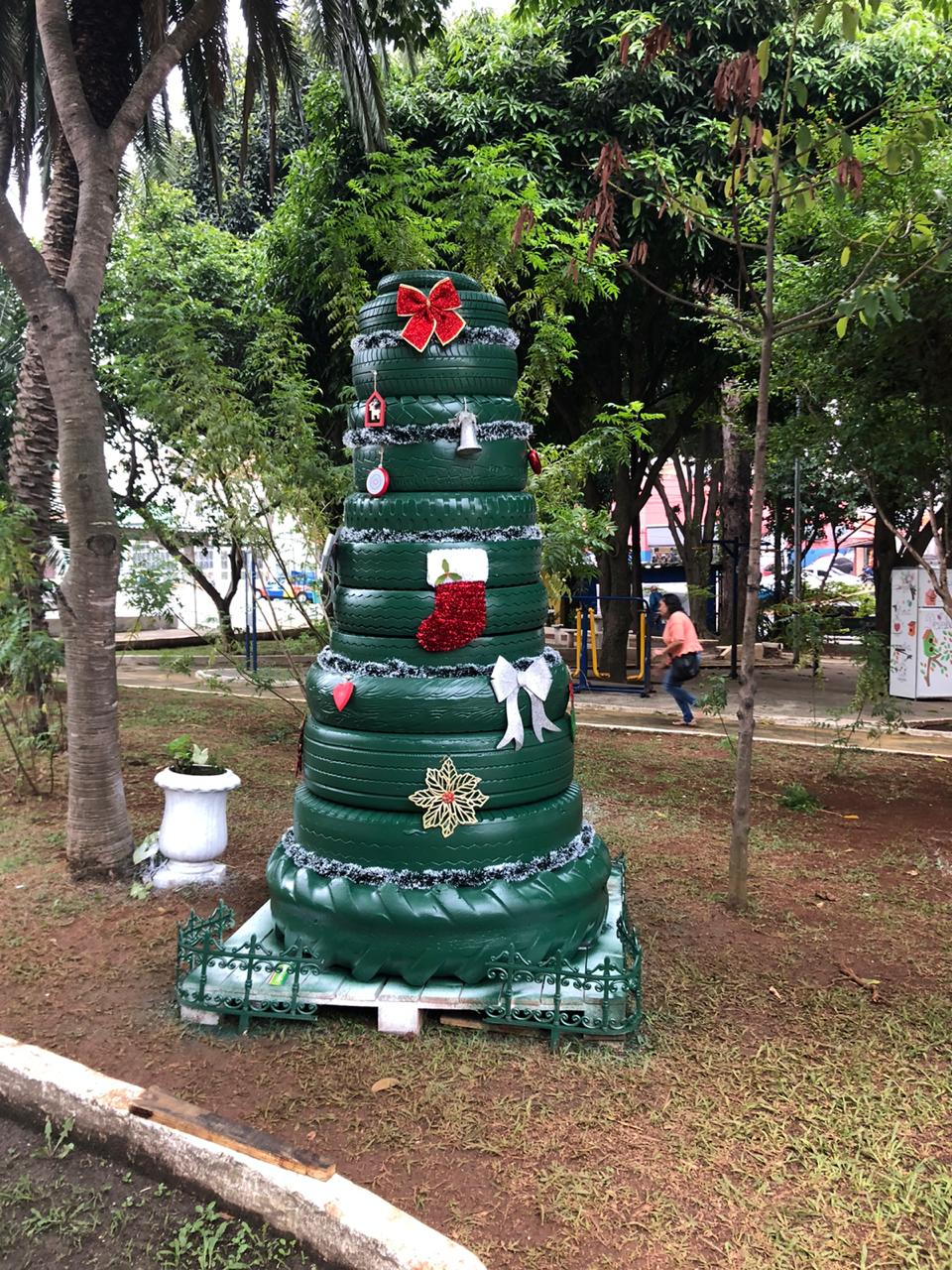 Visualiza-se a imagem de pneus verdes, postos um em cima do outro, simbolizando uma árvore de natal. Há enfeites natalinos colocados na decoração, que está localizada em uma praça.